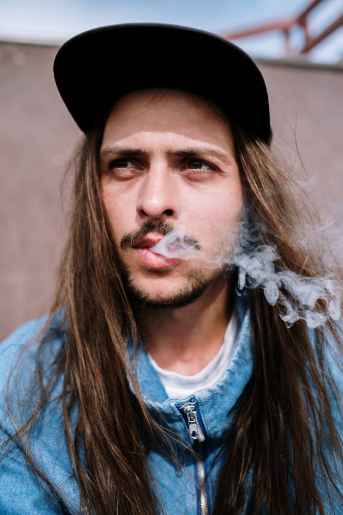 Casual portrait of a young man smoking with long hair and a cap, conveying a relaxed, modern style.