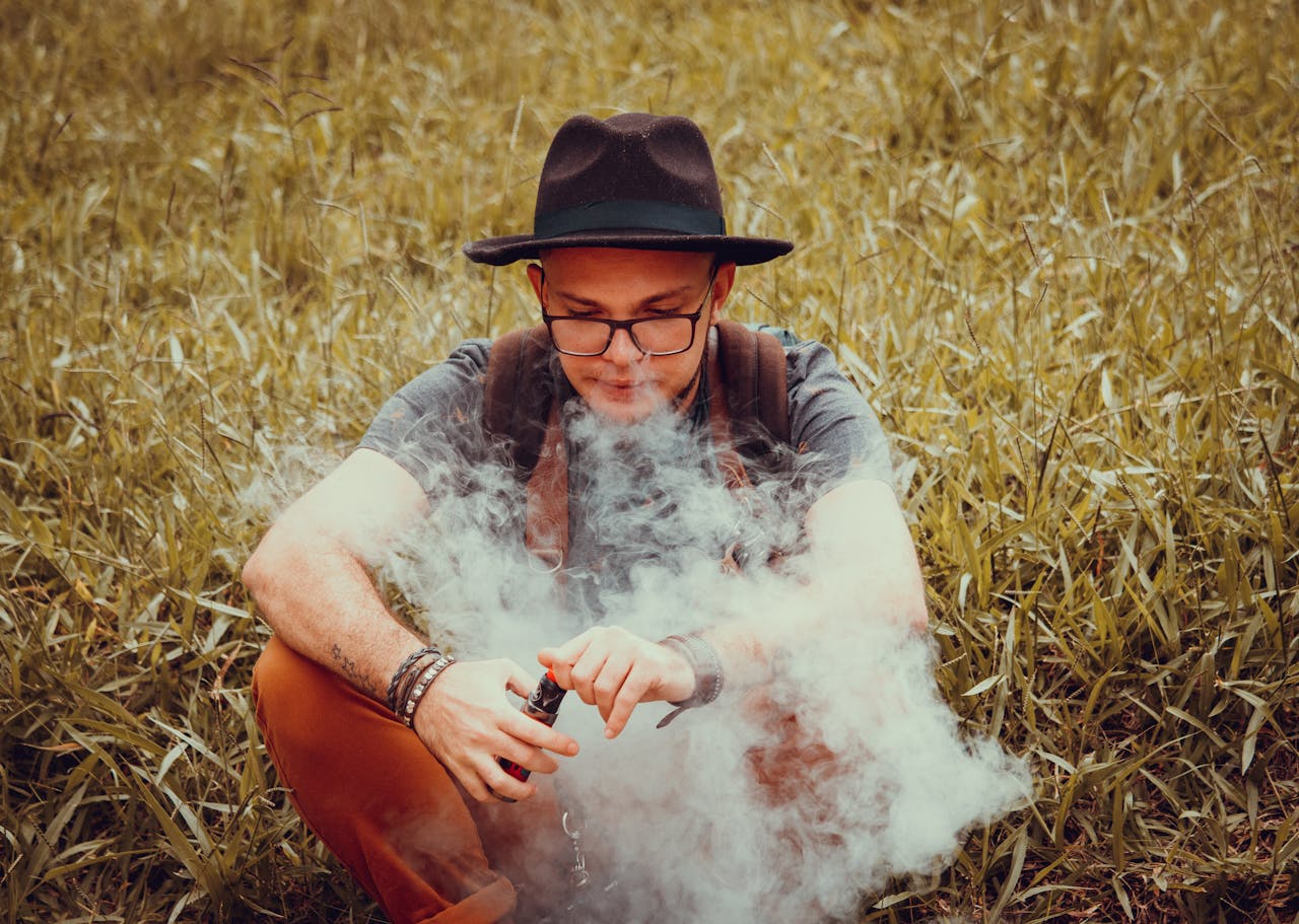 A man with a cowboy hat is sitting in a grass field, vaping on a clear day.