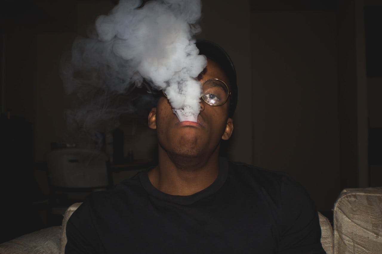 A dark and moody portrait of a man exhaling vape smoke, wearing glasses in an indoor setting.
