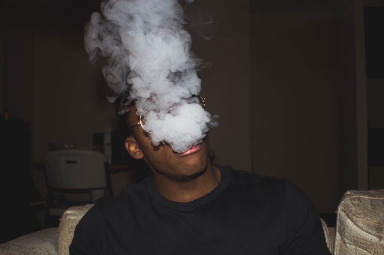 Black man sitting on sofa exhaling thick vapor, captured indoors with moody lighting.