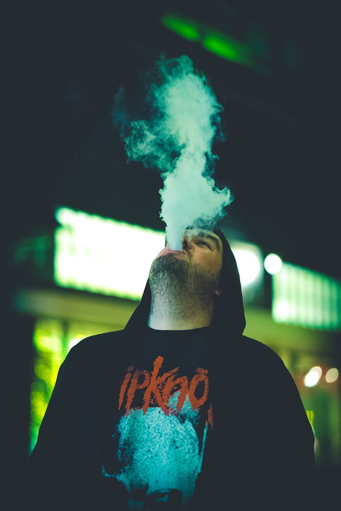 A young man exhales vapor in a neon-lit environment, creating a dramatic ambiance.