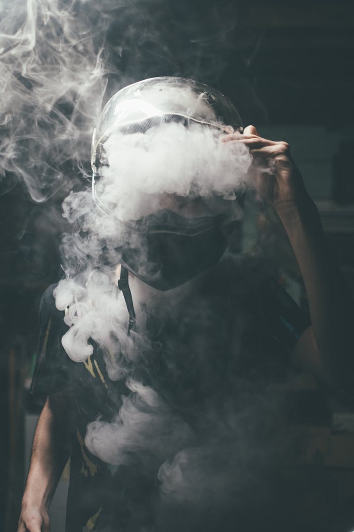 Dramatic portrait of a man wearing a motorcycle helmet surrounded by vape smoke in a dark setting.