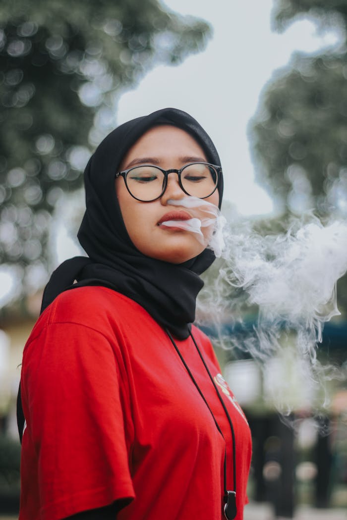 Woman in hijab and red shirt vaping outdoors, exhaling smoke with calm expression.